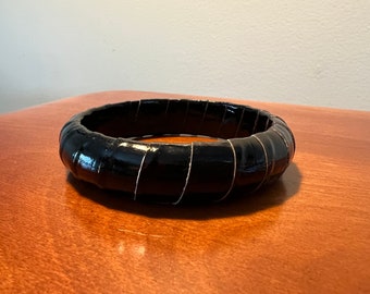 Może przedstawiać: Czarna bransoletka typu bangle z błyszczącym wykończeniem i fakturowanym wzorem. Bransoletka wykonana jest z wielu warstw czarnego materiału, tworząc unikalny i przyciągający wzrok wygląd.
