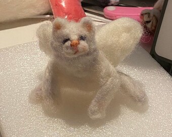 May include: A white felted cat with a fluffy tail, sitting on a white surface.