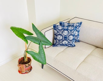 May include: A white couch with two blue and white patterned throw pillows. A green plant in a colorful pot is in the foreground.