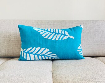 May include: A turquoise blue throw pillow with a white leaf design. The pillow is sitting on a light gray couch.