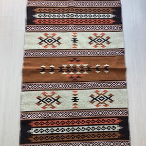 May include: A brown, white and orange patterned rug with fringe on the edges. The rug has a geometric design with a central diamond shape.
