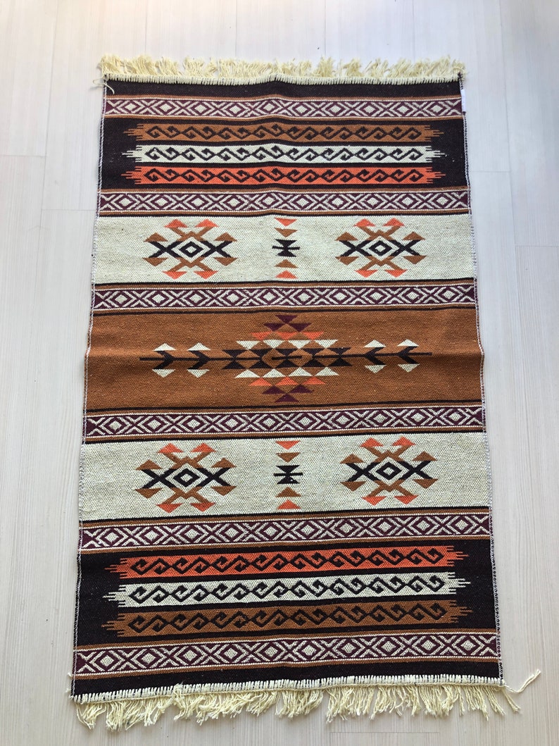May include: A brown, white and orange patterned rug with fringe on the edges. The rug has a geometric design with a central diamond shape.