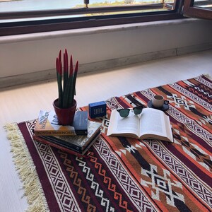 May include: A red and green succulent plant in a terracotta pot sits on a colourful rug with a book, sunglasses, and a small blue speaker. The rug has a geometric pattern in shades of red, orange, brown, and white.