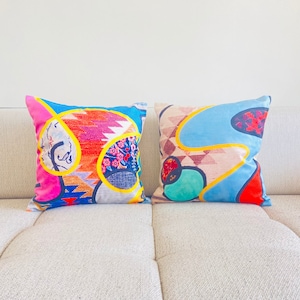 May include: Two colorful throw pillows with abstract geometric patterns in shades of pink, blue, yellow, and green. The pillows are on a white couch.
