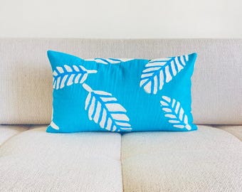 May include: A turquoise blue throw pillow with a white leaf pattern. The pillow is sitting on a light beige couch.