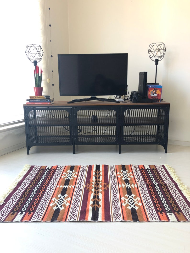 May include: A black metal TV stand with a wood top and wire shelves. A flat screen TV is on top of the stand. There are two black geometric lamps on the stand, one on each side of the TV. A red potted plant is on the left side of the TV. There are books on the stand below the TV. A patterned rug is on the floor in front of the stand.