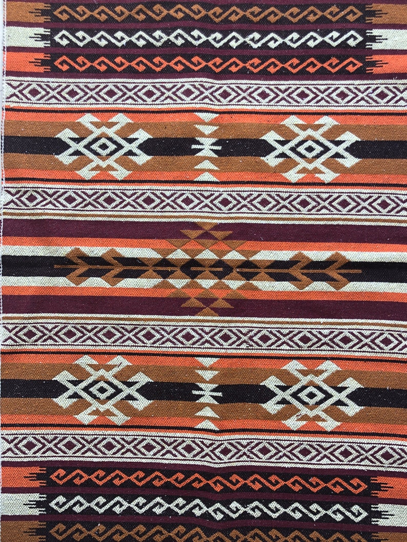 May include: A woven rug featuring a geometric pattern in shades of brown, orange, white, and purple. The rug has a traditional design with repeating motifs.