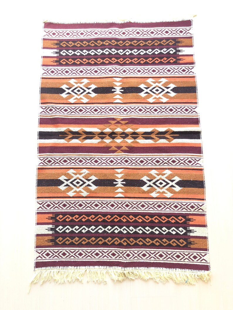 May include: A brown, orange and white woven rug with a geometric pattern. The rug has a fringe on the bottom.
