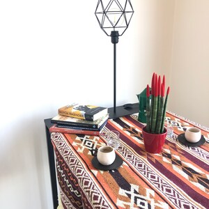 May include: A black geometric lamp with a light bulb sits on a table with a stack of books, a potted plant, and two cups of coffee. The table has a colourful patterned tablecloth with red, orange, brown, and white stripes and geometric designs.