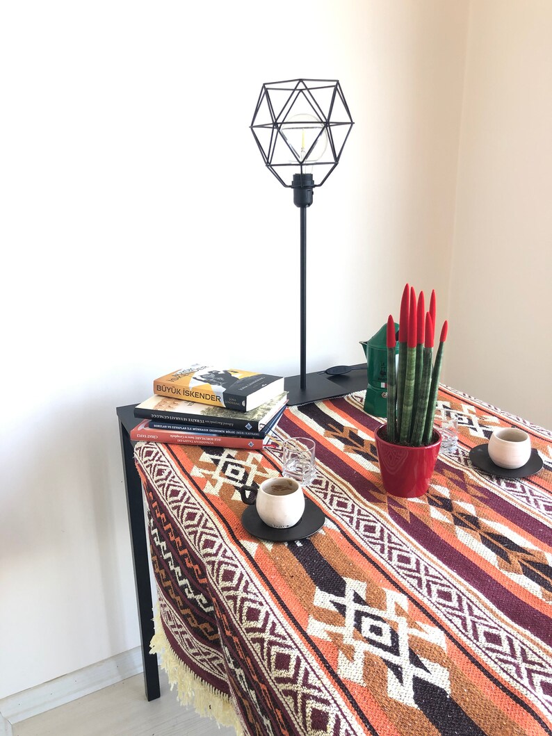 May include: A black geometric lamp with a light bulb sits on a table with a stack of books, a potted plant, and two cups of coffee. The table has a colourful patterned tablecloth with red, orange, brown, and white stripes and geometric designs.