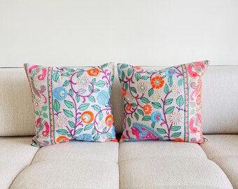 May include: Two decorative throw pillows with a floral pattern in shades of pink, blue, green, orange, and white. The pillows are on a light beige couch.