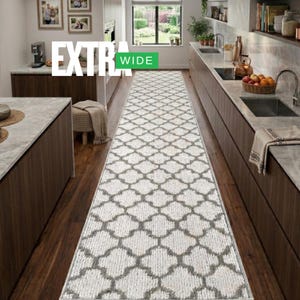 May include: A long, rectangular runner rug featuring a repeating white and gray quatrefoil pattern. The rug is positioned on a dark wood floor between two kitchen counters. A green rectangular label with the white text EXTRA WIDE is visible.