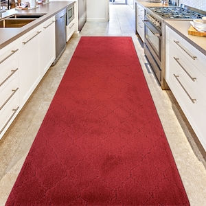 May include: A red runner rug with a geometric pattern in a kitchen setting. The rug is placed between white kitchen cabinets.