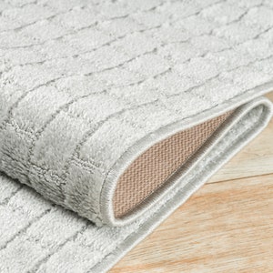 May include: Close-up of a gray area rug with a textured woven pattern. The rug is folded over, showing the beige backing.