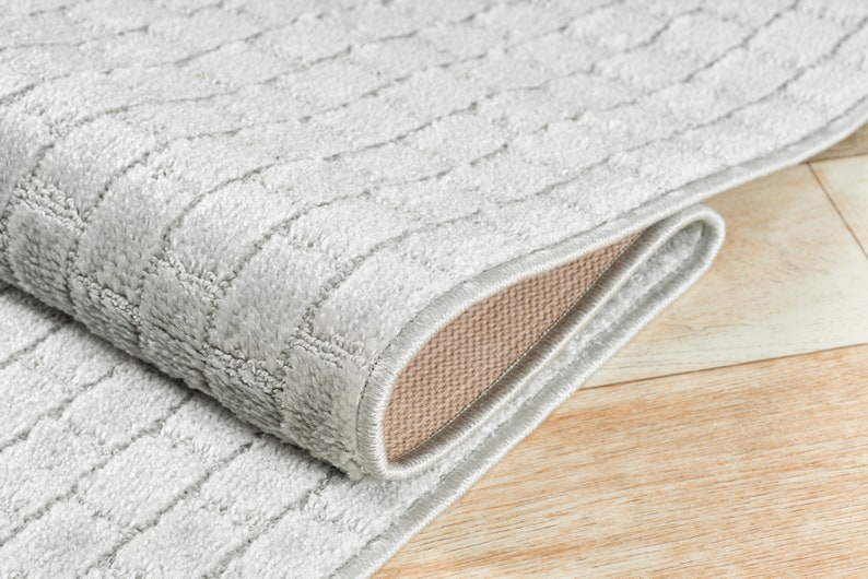 May include: Close-up of a gray area rug with a textured woven pattern. The rug is folded over, showing the beige backing.