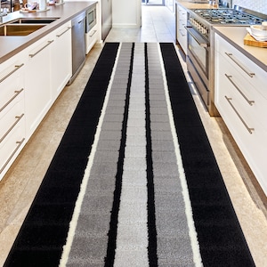 May include: A black and gray striped runner rug with white accents. The rug is placed in a kitchen hallway between two white cabinets.