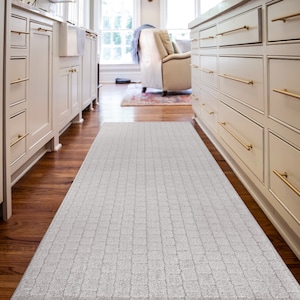 May include: A white kitchen runner rug with a textured, checkered pattern. The rug is placed between two sets of white kitchen cabinets with gold hardware.