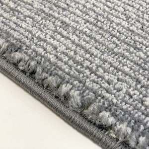 May include: Close-up of a gray rug with a textured surface and a stitched edge.