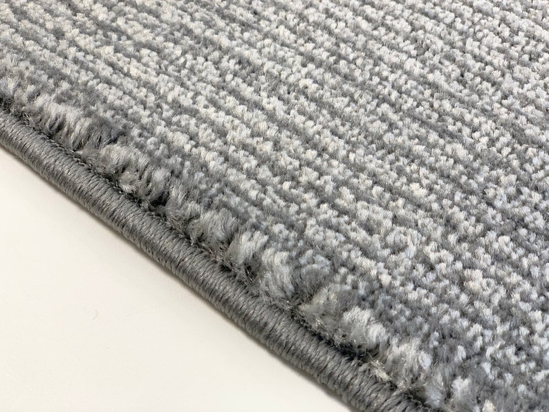 May include: Close-up of a gray rug with a textured surface and a stitched edge.