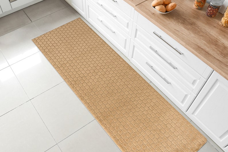May include: A beige kitchen runner rug with a textured woven pattern. The rug is placed in front of white kitchen cabinets with silver handles.