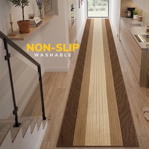 May include: A long, rectangular runner rug featuring a striped pattern in shades of brown, tan, and cream. The rug has a textured surface with darker brown edges and a lighter central stripe. The text NON-SLIP WASHABLE is printed in yellow and white.