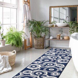 May include: A white bathroom with a white bathtub, a wooden vanity with a white sink, and a blue and white patterned bath rug. The bathroom has a large window with plants on the windowsill and a white robe hanging on a wooden ladder.