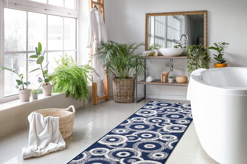 May include: A white bathroom with a white bathtub, a wooden vanity with a white sink, and a blue and white patterned bath rug. The bathroom has a large window with plants on the windowsill and a white robe hanging on a wooden ladder.