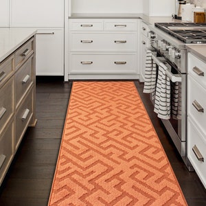 May include: A kitchen with white cabinets and a brown kitchen island. A patterned orange runner rug is placed on the floor between the cabinets and the island.