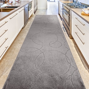 May include: A gray runner rug with a repeating circular pattern. The rug is rectangular and placed in a kitchen between white cabinets and appliances. The rug's design features overlapping circles in a lighter shade of gray.