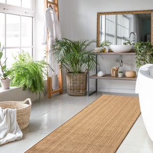 May include: A bathroom with a white bathtub, a brown woven rug, and a large window with plants. The bathroom has a white sink and a mirror with a gold frame.