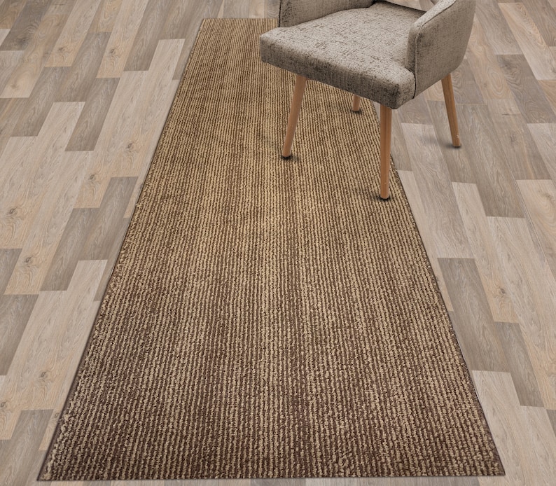 May include: A brown and beige striped runner rug with a subtle textured pattern. The rug is placed on a light wood floor.