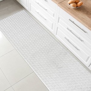 May include: White kitchen runner rug with a textured woven design. The rug is placed in front of white kitchen cabinets with silver handles.