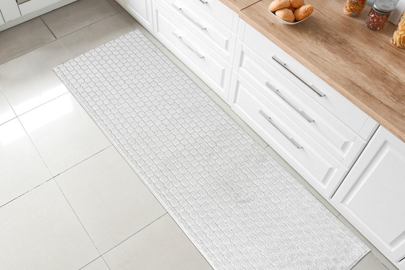 May include: White kitchen runner rug with a textured woven design. The rug is placed in front of white kitchen cabinets with silver handles.