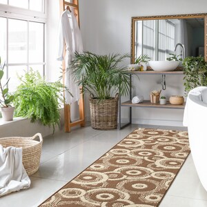 May include: A brown and cream patterned bath rug with a circular design. The rug is placed in a bathroom with a white bathtub, a wooden vanity with a sink, and plants.