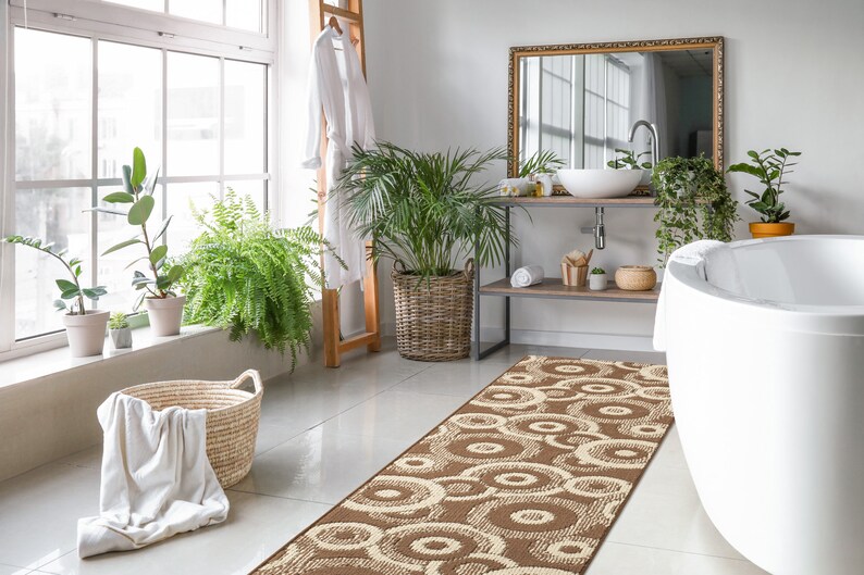 May include: A brown and cream patterned bath rug with a circular design. The rug is placed in a bathroom with a white bathtub, a wooden vanity with a sink, and plants.