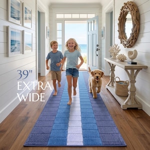May include: A rectangular runner rug featuring a gradient of blue and white blocks with a central white stripe. The rug is labeled 39" EXTRA WIDE, indicating its width. It is positioned on a wooden floor in a hallway, showcasing a textured surface.