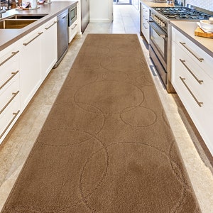 May include: A brown kitchen runner rug with a circular pattern. The rug is placed between two white kitchen cabinets.