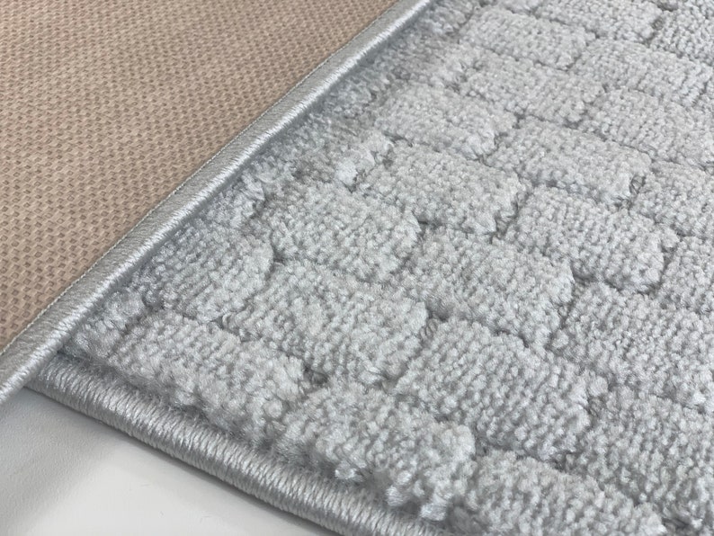 May include: Close-up of a gray bath mat with a textured surface and a beige backing. The mat has a stitched edge.