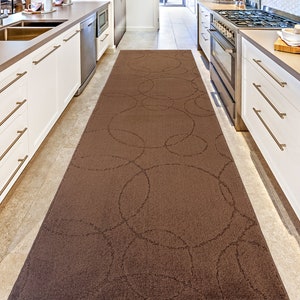 May include: A brown kitchen runner rug with a circular pattern. The rug is placed in a kitchen with white cabinets and a stainless steel oven.