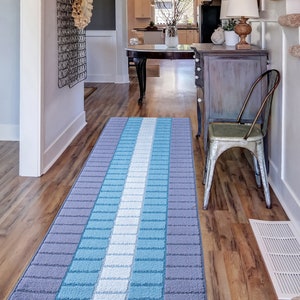 May include: A blue and gray striped runner rug with a white stripe down the center. The rug is placed in a hallway with hardwood floors and a wooden staircase.
