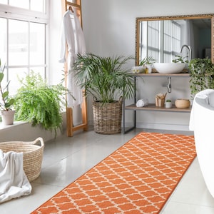 May include: A bathroom with a white bathtub, a brown wooden vanity with a white sink, and a large orange and white patterned rug. There are plants in pots and a white towel hanging on a ladder.