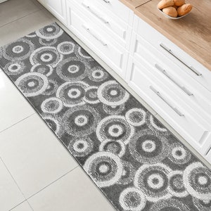May include: A gray and white patterned runner rug with a circular design. The rug is placed in front of a white kitchen cabinet.