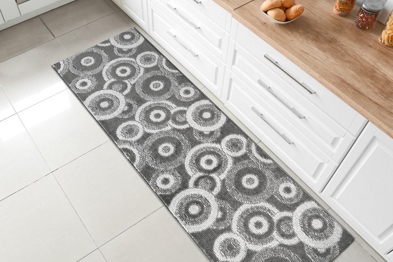 May include: A gray and white patterned runner rug with a circular design. The rug is placed in front of a white kitchen cabinet.