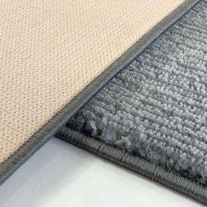 May include: Close-up of the edge of two rugs. One rug is a light beige color with a woven texture. The other rug is a dark gray color with a fuzzy texture. Both rugs have a dark gray border.