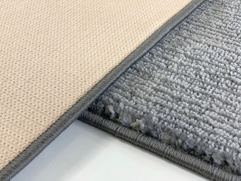 May include: Close-up of the edge of two rugs. One rug is a light beige color with a woven texture. The other rug is a dark gray color with a fuzzy texture. Both rugs have a dark gray border.