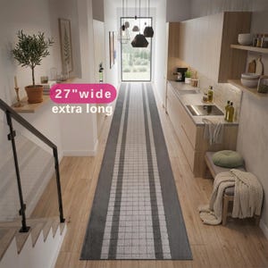 May include: An extra long runner rug with a 27-inch width, featuring a central grid pattern in light gray and white, bordered by solid dark gray stripes. The rug is positioned on a light wood floor in a narrow hallway between a kitchen and stairs.