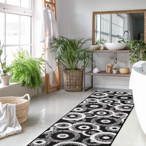 May include: A black and white bathroom rug with a circular pattern. The rug is placed in front of a white bathtub and a wooden shelf with a sink and mirror.