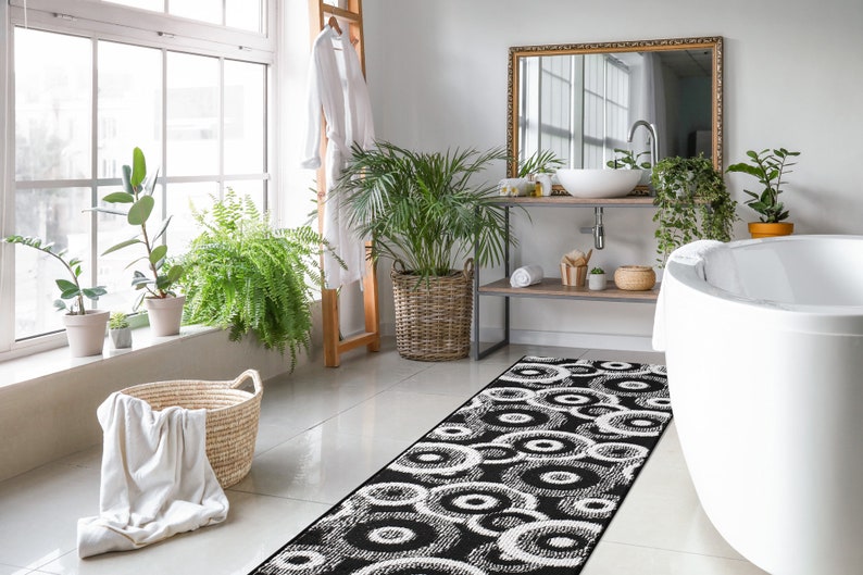 May include: A black and white bathroom rug with a circular pattern. The rug is placed in front of a white bathtub and a wooden shelf with a sink and mirror.