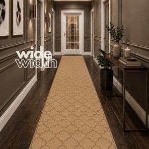 May include: A long, tan runner rug featuring a repeating quatrefoil pattern in a darker shade. The rug is positioned centrally along a dark wood floor in a hallway, with the text "wide width" displayed in white letters on the left side.
