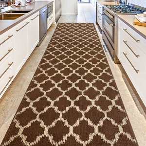 May include: A brown and cream patterned runner rug with a geometric design. The rug is placed in a kitchen hallway between white cabinets.